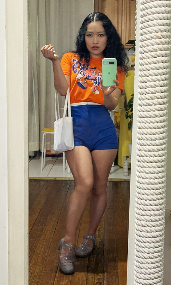 Reflection in the mirror is showing Nana wearing the Knicks T-shirt, cobalt blue shorts, and the mini tote